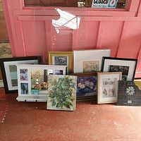 Overview of the lot showing 9 various framed art pieces including prints in multiple frame styles and a floral wood mirror leaning against a pink wall