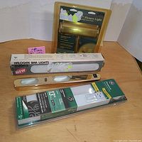 Overview photo showing all four lighting fixtures in lot including the brass picture light, halogen bar light, brass fluorescent fixture, and packaged bulb.