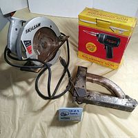 Photo of all three tools together on a surface: Skilsaw power saw, soldering gun box, and antique hand stapler