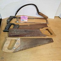 Three vintage hand saws and one grass/hay sickle arranged on a wooden surface showing handles and blades.