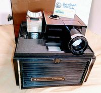 Front and top view of the Bell & Howell Slide Cube Projector showing the cube slide holder with multiple slides and the large projector lens with woodgrain detail on top.