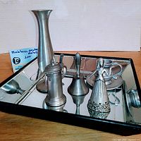 Five vintage metal miniature collectibles arranged on a mirrored tray including a tall Leonard vase, a tankard style miniature pitcher with lid, small bell with handle, figurine of a woman in dress, and a small rounded pitcher with handle.