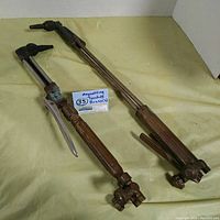 Two vintage oxy acetylene cutting torches laid on a beige surface showing their full length and overall condition.