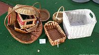Photo showing various wicker baskets stacked and grouped on a green carpeted floor. Includes large oval basket, white rectangular basket, rectangular basket with black weave, and others.