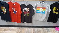 Front view of five video game theme t-shirts hung on a wall, including Super Mario Kart, Mario Brothers, PlayStation, and Pac-Man graphics.