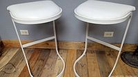 Two white vinyl padded Mid-Century Modern bar stools with white tubular metal frames, placed on wood floor against a gray wall.