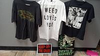 Full view of all three T-shirts hanging above three marijuana-themed signs on floor, showing lot contents