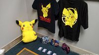 Large yellow Pikachu plush seated on floor next to two black Pikachu-themed T-shirts hanging on wall, two blue and one pink Pokémon collector tins on floor, and five Pokémon cards spread out in front