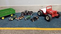 Photo showing a lineup of seven vintage metal toy vehicles including a John Deere trailer, Tonka dune buggy, Jeep labeled Ruger, Coca-Cola tow truck, and four small vintage cars.
