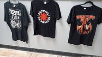 Three black rock band t-shirts hung on hangers against a wall: My Chemical Romance, Red Hot Chili Peppers, and Twenty One Pilots.