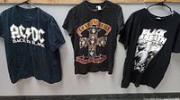 Front view of three black rock band T-shirts: AC/DC, Guns N' Roses, Black Sabbath hung on hangers.