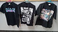 Three black graphic T-shirts hanging on hangers showing Miami Vice, Scarface, and Las Vegas prints