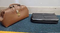 Brown leather doctor purse and black travel kit bag on carpeted floor against white wall