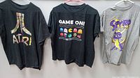 All three t-shirts hanging on hangers against a wall: Atari black, Pac-Man black, Spyro gray.