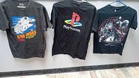Three graphic T-shirts hung on hangers showing Star Wars, PlayStation, and Mortal Kombat X designs.