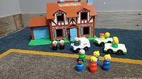 Front view of the vintage Fisher Price play house with all figures and vehicles arranged in front of it on carpet.