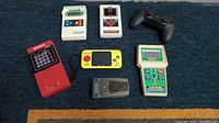 Overview of all seven handheld electronic games and devices arranged on carpet, showing size and condition.