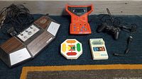 Top-down photo showing five vintage electronic gaming items including a pong TV game console, orange Replay Copycat handheld game, Bambino Basketball handheld, Mattel Electronics Football game, and Sony PlayStation controller.