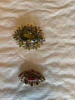 Two antique brooches on fabric background showing larger green gem brooch and smaller reddish gem brooch