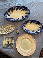 Lot displayed on grey fabric, shows two blue floral platters, oval floral plate, small egg cup, Delft hot plate and Royal Doulton plate