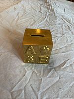Angled view of the brass cube child's bank showing the embossed letter A, B and animal motifs on its side, with coin slot on top.