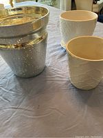 Three planters displayed on a blue cloth: large silver speckled planter and two white ceramic planters with textured surfaces.