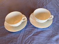 Front view of two white embossed leaf pattern teacup and saucer sets laid on dark surface.