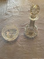 Crystal decanter with stopper and three-footed crystal bowl placed on fabric surface.
