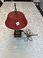 Full view of the decorative table lamp showing red metal shade, brass finial, brass stem with detailing and square wooden base, with cord and plug laid out beside it