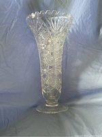 Front view of the tall clear crystal vase with deep hand cut floral and geometric patterns, showing the notched rim and circular foot base on a blue background.