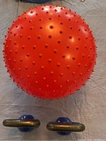 Red textured fitness exercise ball with small spikes and two blue kettlebells lying below it on a cloth surface, taken from above.