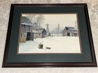 Full view of the framed winter farmyard scene print depicting barns, windmill, and snow with chopping block in foreground.