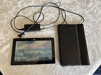 Photo showing the Microsoft Surface tablet powered on with screen on, the protective black cover, and the two charger power adapters with cords, all laid out on a table.