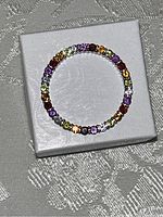 Top-down view showing circular ring band with colorful gemstones set around the band on a white textured box.