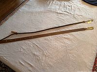 Two brown leather belts laid out on a cream fabric surface, showing full length and buckle ends with attached tag on one belt.