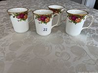 Four Royal Albert Old Country Rose coffee mugs arranged on a patterned tablecloth, showing front view of floral and gold rim details.