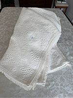 White cotton single twin bedspread with diamond and square textured pattern laid on a floral bedspread. Shows fringe trim on the edges.