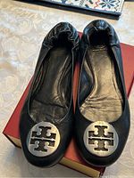 Pair of black leather Tory Burch ballet flats with silver metal disc logo on toe caps, displayed on a red box.