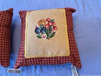 Two petit point pillows photographed on a blue surface, showing red checkered borders and stitched floral designs on beige fabric centers.