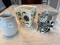 Four flower containers on a table: white ceramic vase, square ceramic floral planter, ceramic cat-shaped container, and silver metal square container.