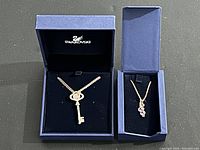 Two Swarovski necklaces displayed in their blue branded boxes on a black background; left box contains the key pendant necklace, right box contains the necklace with three crystal pendant.