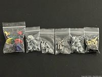 Six individual small plastic bags each containing different sets of Monopoly game pieces including metal tokens and colorful plastic figurines.
