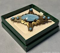 Overall view of the brooch in its box showing the brass design, central blue stone, and blue enamel accents.