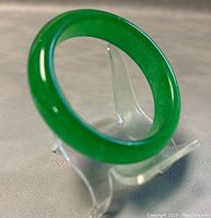 Polished green jade bangle displayed upright on clear stand showcasing smooth and glossy finish.