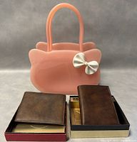 Front view of pink jelly kitty cat purse with white bow and two vintage brown Rolfs wallets on box lids in front of purse.