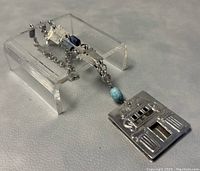 Side-angle photo showing the pendant and part of silver chain laid on a clear acrylic stand, highlighting the blue stone bead and chain design with chain links and decorative shaped silver elements.