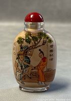 Front view of snuff bottle showing a man and a woman in traditional attire with calligraphy and tree details, capped with a red stopper.