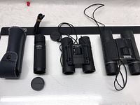 Lot of three optic devices including black Bushnell 10x25 binoculars, Bushmaster 8x21 binoculars, and Pentax 7x20 monocular with protective case and lens cap.