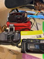 Three vintage film cameras including Pentax AF Zoom black camera, Kodak Cameo Motor EX gray model, Ricoh FF-70d black with manual, on tabletop with red camera bag.