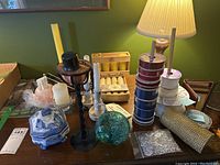 Overview of natural beeswax candles, black metal candleholder, wooden box with candles, multiple ribbon spools, handcrafted glass ornament, blue-and-white ceramic candle pot, some candles, and additional items on a wooden table.